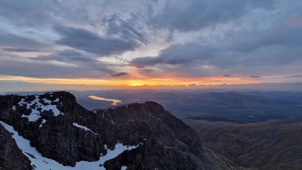 The sun sets behind mountains, casting warm colours over the valley below.